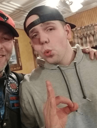 Michael Moura in a hoodie and backwards baseball hat makes the "okay" hand gesture. Appears to be in a function hall or restaurant and posing with another man in a leather jacket who is mostly off camera.
