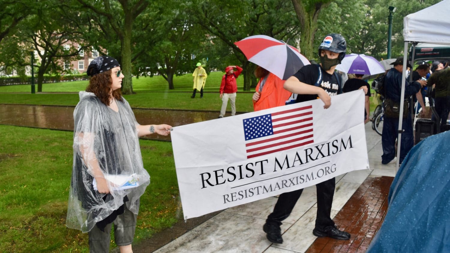 Photo of a gathering of people in a park. Two white people in foreground, one in helmet and face mask, hold up a banner with United States flag and the text "Resist Marxism, resistmarxism.org."