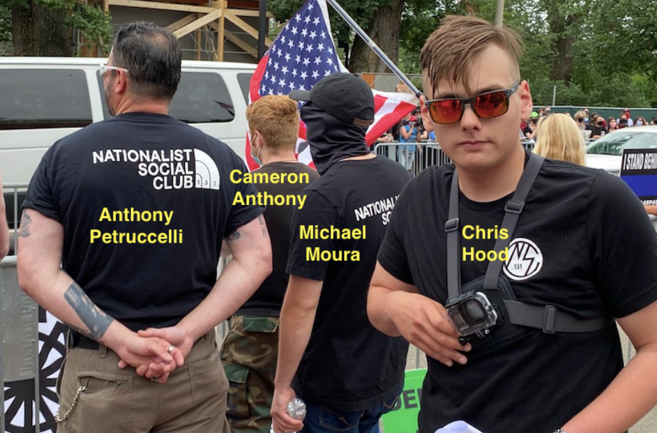 Photograph of man in sunglasses, a Nationalist Social Club shirt, and with a Go Pro strapped to his chest facing the camera and 3 men dressed in black facing away. Man facing the camera is labelled Chris Hood and other 3 are labelled, left to right, Anthony Petruccelli, Cameron Anthony, and Michael Moura. Petruccelli wears a shirt that reads "Nationalist Social Club" next to what looks like a North Face logo. Police barricades and a crowd are in the background.