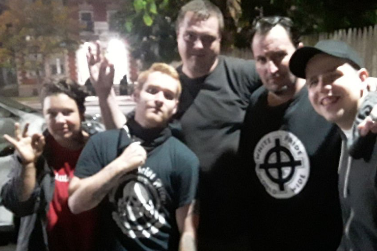 Blurry group photo of 5 white men, mostly dressed in black, at night. Two making the "okay" hand gesture. One has a shirt that reads "white pride world wide" and another in a skull shirt that appears to read "Patriot Front"
