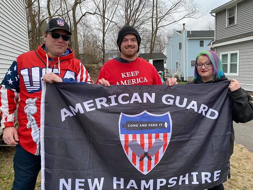 Photo of John Camden in "USA" hoodie and two other people, one in Trump shirt stand outside holding up flag reading "American Guard New Hampshire" with the American Guard logo. That log is a red white and blue shield with a cannon reading "com and take it" and two crossed butcher's cleavers.