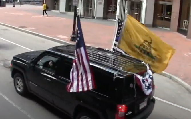 Black SUV on city street with large flags attached to it. Betsey Ross US flag on the left, Gadsen and thin blue line US flags on the right, and semicircular US flag banner on the back.