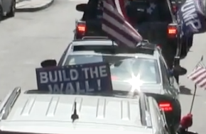 Photo of line of cars in a procession, lots of US flags visible long with a Trump flag and a "Build the Wall" sign.