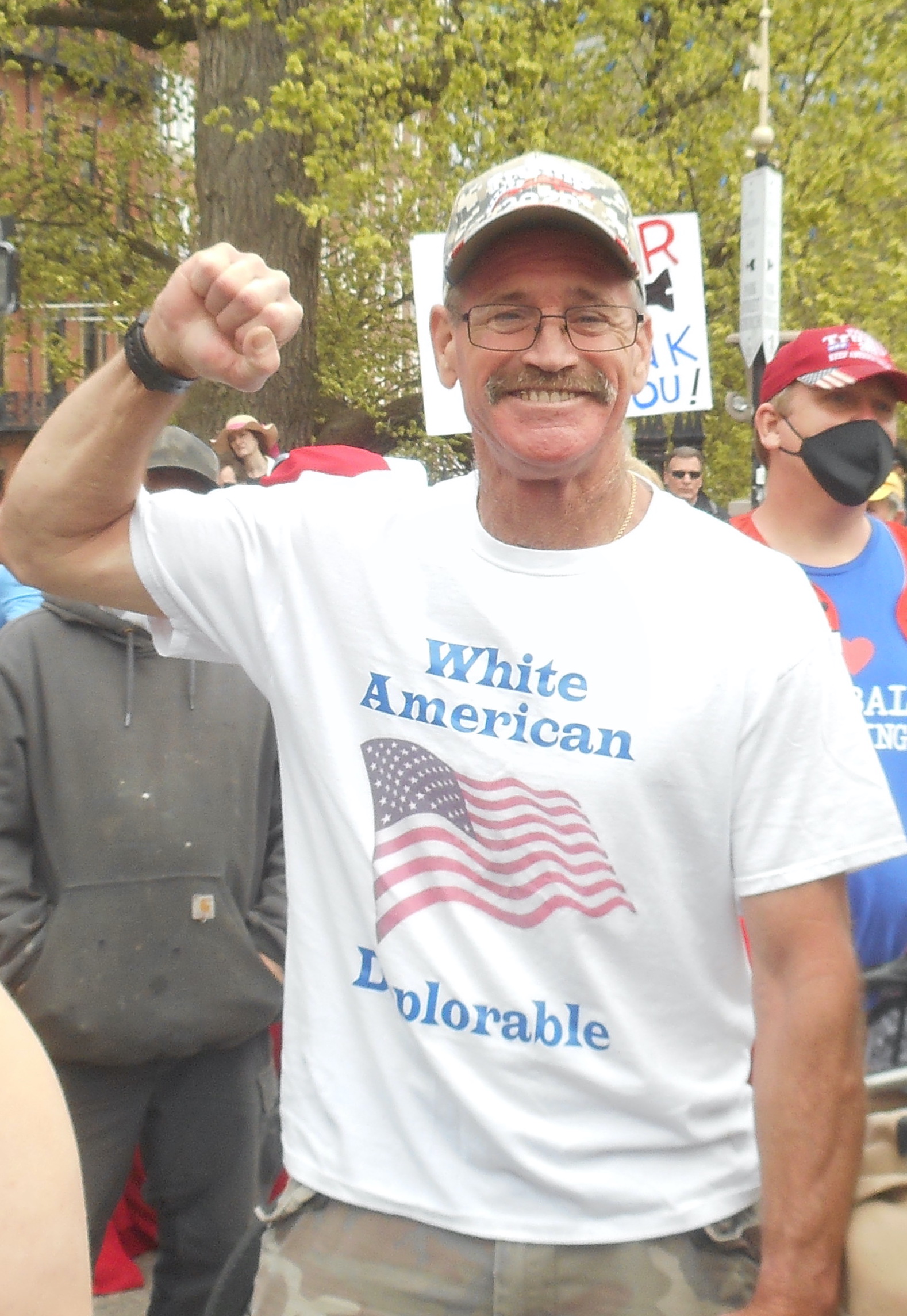 Man with mustache and classes raises fist at a rally. White shirt has a US flag an dreads "White American Deplorable"