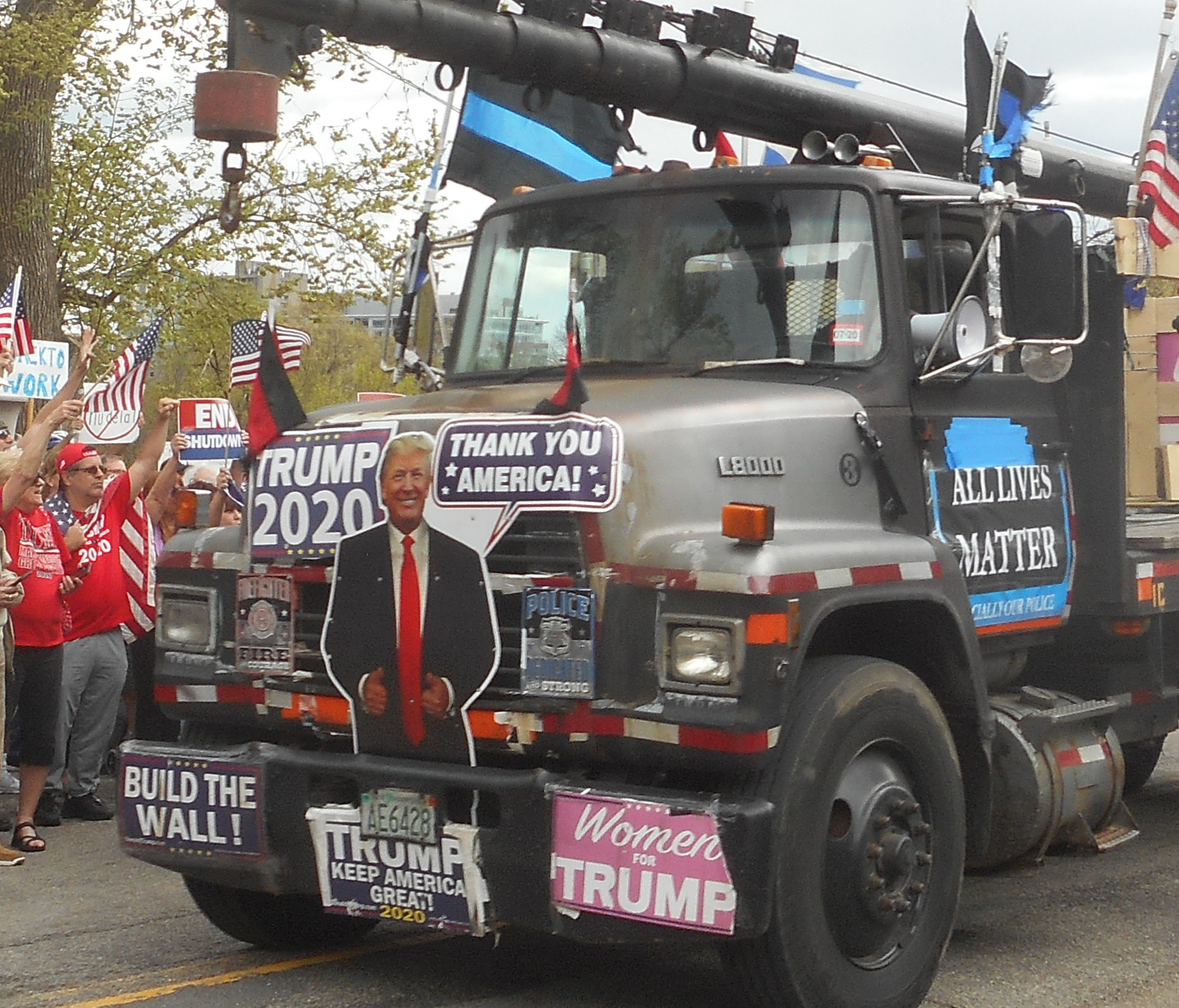 Photo of large black truck with crane. Crowd in red with US flags raise their fists at in in the background. Truck is coerd in Trump signs including a cutout of Trump, "Build the Wall," "Trump 2020" "Women for Trump," "All Lives Matter," and thin blue line flags.