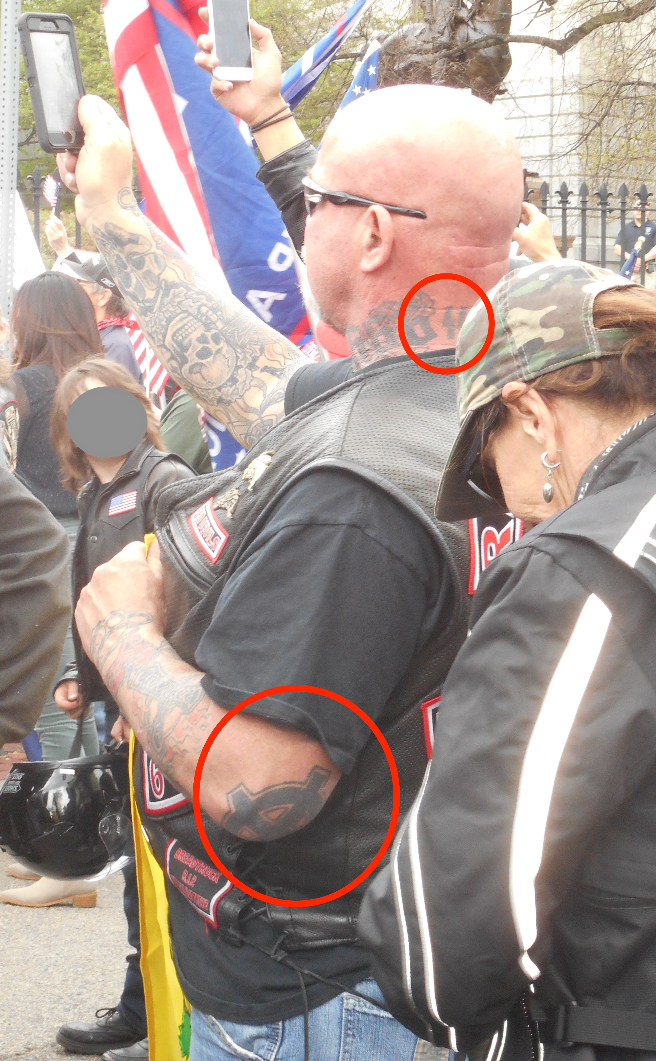 Bald man with leather biker vest at a rally. Man has many tattoos and red circles point out a Nazi "SS" bolt neck tattoo and the white supremacist version of the rounded Celtic cross on his left elbow.