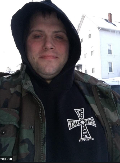 Headshot of John Camden (Josh Cameron). He is wearing a camouflage jacket and looking into the camera, with a slight close-lipped smile. Underneath his jacket he is wearing a black sweatshirt with a large cross on the right-hand side (his left chest area) that says 'White Pride'.