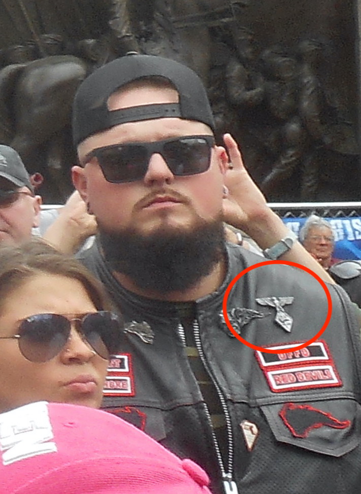 Beared man in a black baseball hat an suglasses in a crowd. Leather jacket has a Nazi eagle emblem pin on it, highlighted by a red circle.