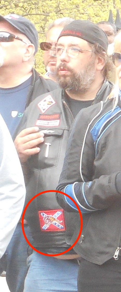 Image of a beared man in a crowd with a leather jacket with a Confederate flag patch on it. Patch is indicated with red circle.