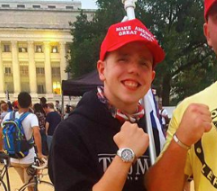 Photo of Michael Moura in a Make America Great Again Hat, holding a flag over their shoulder. They are holding up a fist to micmick the pose of the person next to them whose face is not shown. They are in a public space with people in the background.