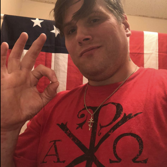 Picture of John Camden in front of a US flag making the "okay" hand gesture. Wears a cross necklace