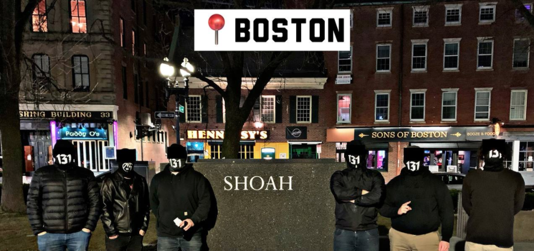 Image of 6 men standing at the New England Holocaust Memorial in Boston at night. They're all wearing face coverings that have the number 131 on them. The piece of the monument they are standing next to reads: "SHOAH" 