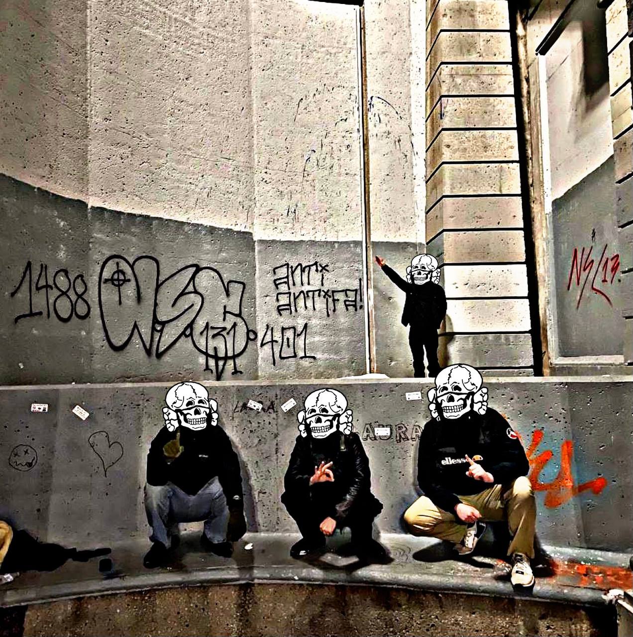 Photo of three people crouching underneath a graffiti wall with 1488 and NSC 131 on it. They are wearing skeleton masks/helmets. Behind them a person, also masked, is doing the Nazi salute. 