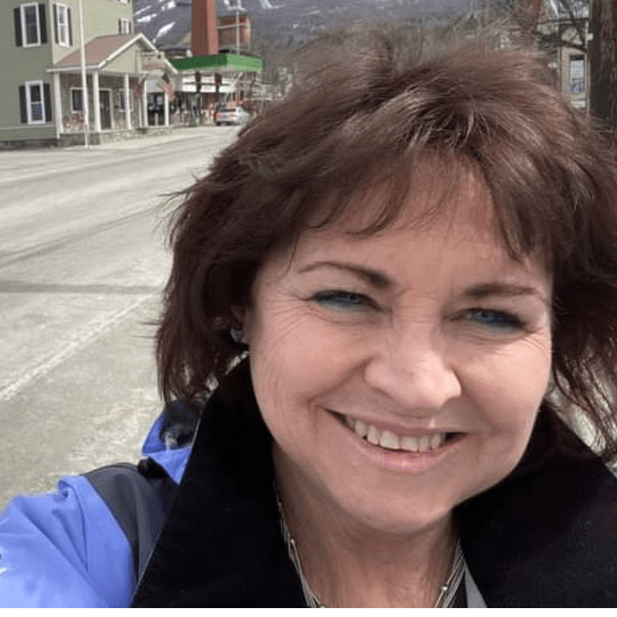 Image of Sue Ianni, a middle-aged white woman with chin-length brown hair and blue eyes. She is smiling and wearing blue eyeliner and a blue jacket. She is standing on a street and appears to be taking a selfie.