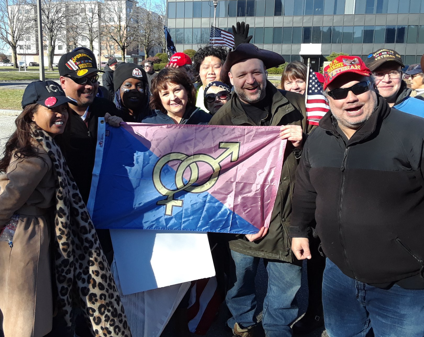 Sue Ianni holds a straight pride flag. She is standing between Mark Sahady and John Hugo who is wearing his tri-cornered hat. Around 10 people are posing for the photo.