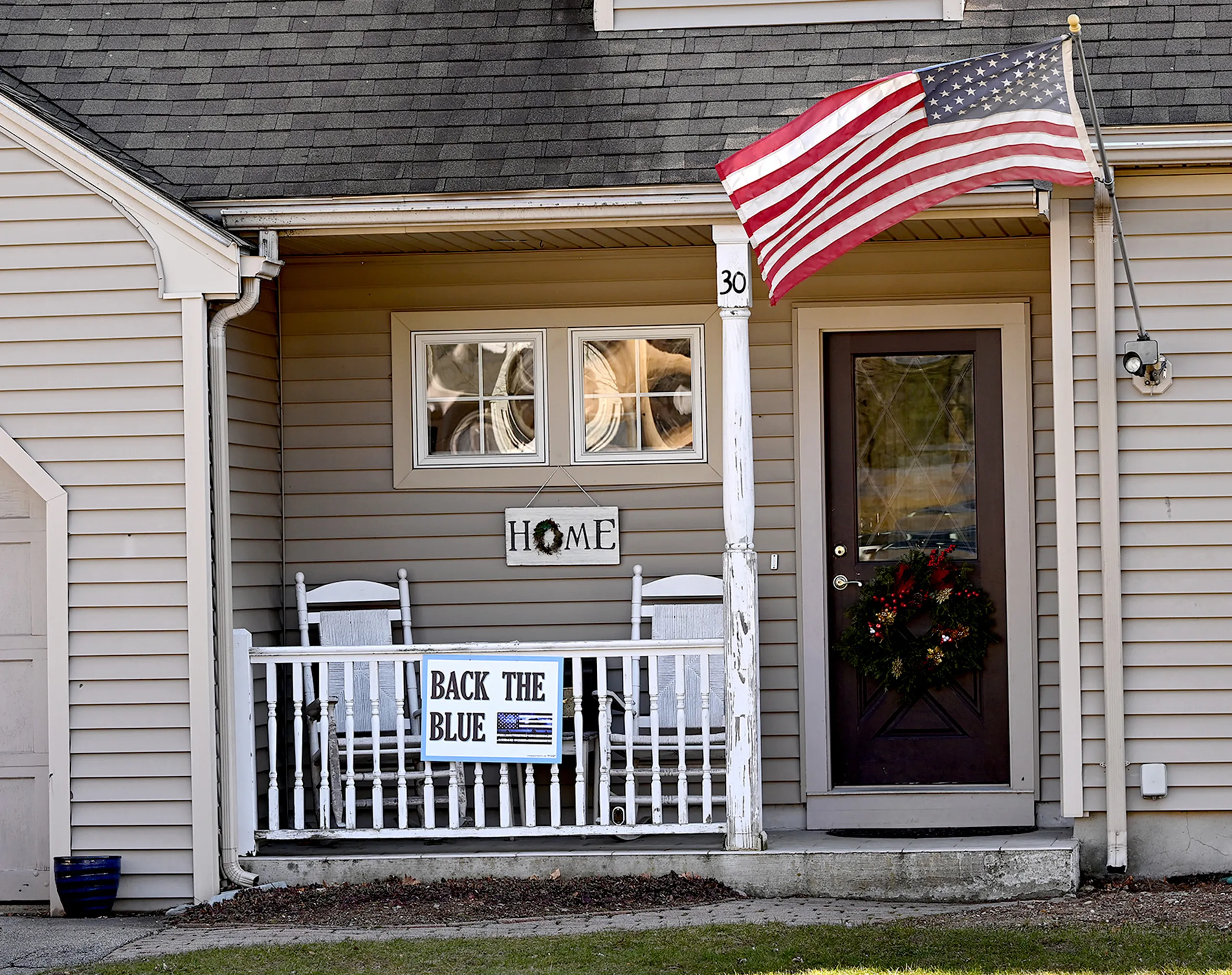 Image of a house. It is painted tan and has the number 30 on it. A Christmas wreath hangs on the door. The porch has two white chairs on it and a sign that says "Home." An American flag and a Back the Blue sign hang on the front of the house.