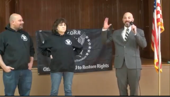 Mark Sahady, Sui Ianna, and Samson Racciopi standing at the front of an auditorium. Sahady and Iannai are wearing CORR hoodies, and Racciopi is wearing a grey suit. Racciopi is holding a microphone and appears to be talking. There is an American flag at the far right of the image, and in the background there is a large black and white CORR banner.