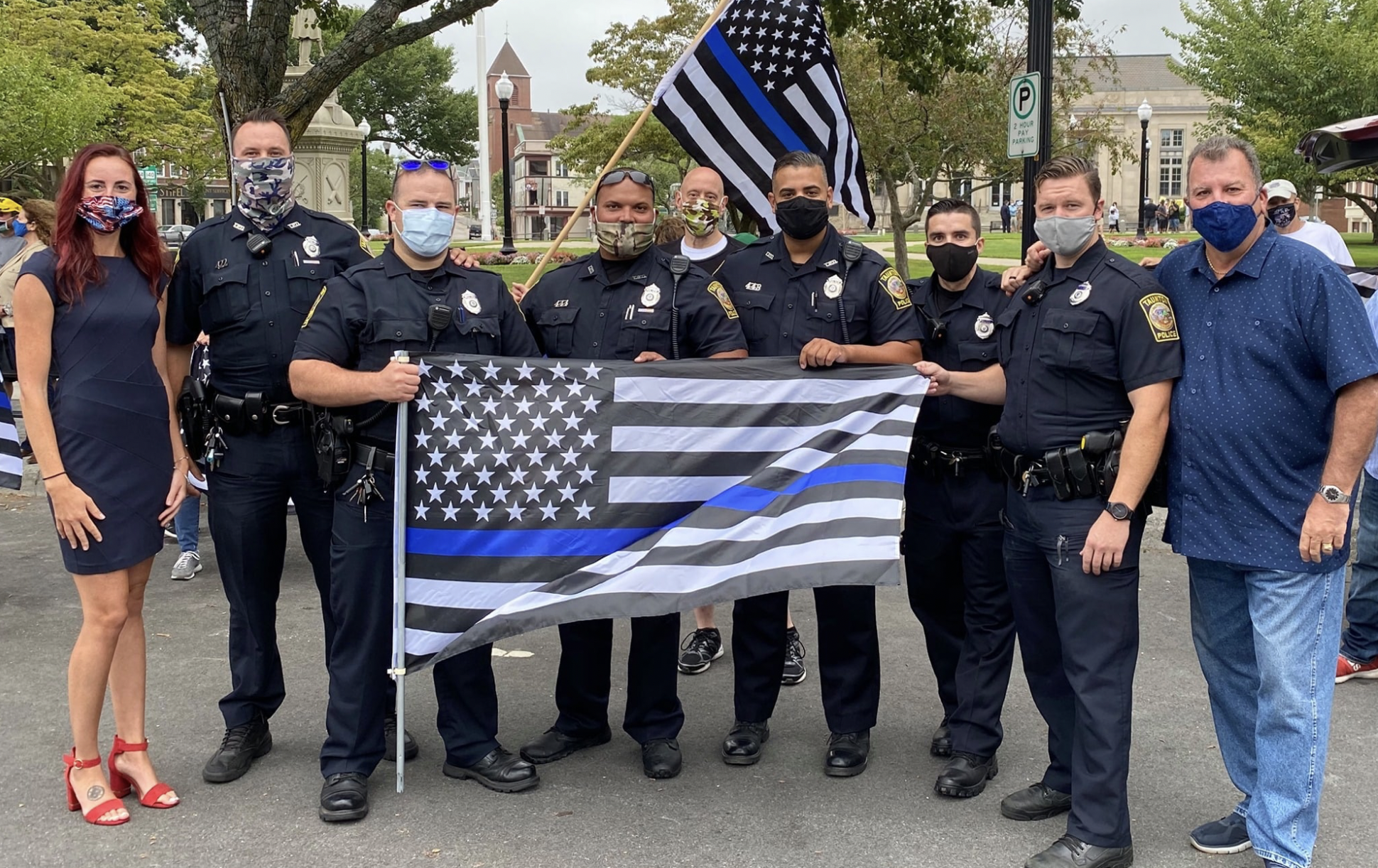 McCaul stands with seven uniformed police officers and a person wearing a navy blue dress and red high heels. They are all wearing face masks. The police hold two large Thin Blue Line flags (a black and white American flag with one stripe colored dark blue). They are outside in a park.