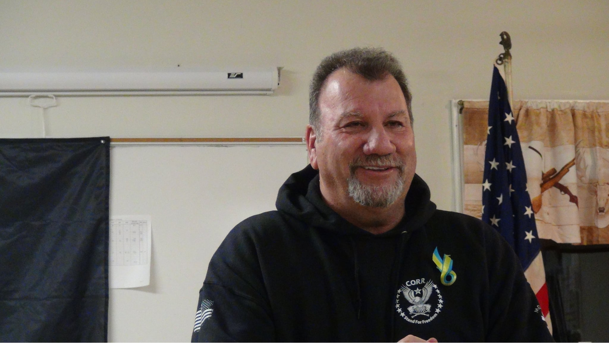 John McCaul stands in front of an American flag. He is wearing a black sweatshirt with the CORR logo and a blue and yellow ribbon pinned to his chest.
