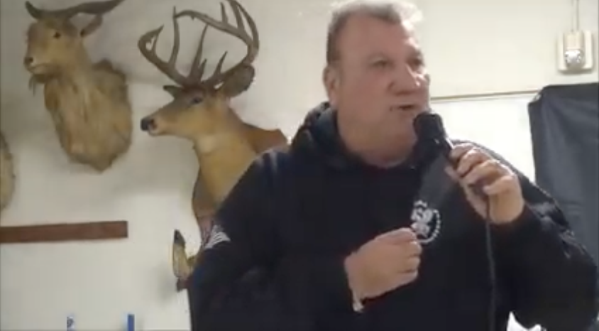John McCaul is speaking into a microphone. His beard is shaved off. He is wearing a black sweatshirt with the CORR logo and indicating the logo with his right hand. Behind him are the heads of two taxidermied animals mounted on the wall.