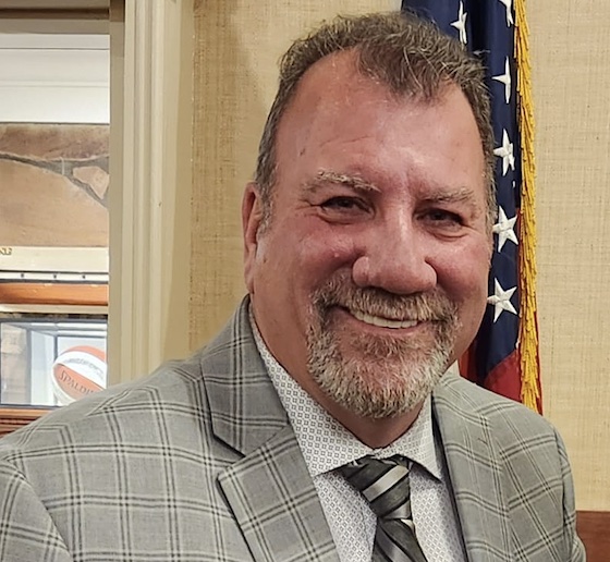 John McCaul stands in front of an American flag. He is a middle-aged white man with salt-and-pepper hair, eyebrows, and beard. He is wearing a gray suit and tie.