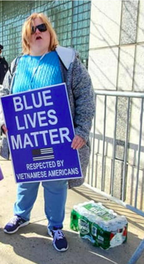 Patenaude, a white woman with chin length blonde hair, stands holding a "Blue Lives Matter" sign. She has a blue sweater and sunglases
