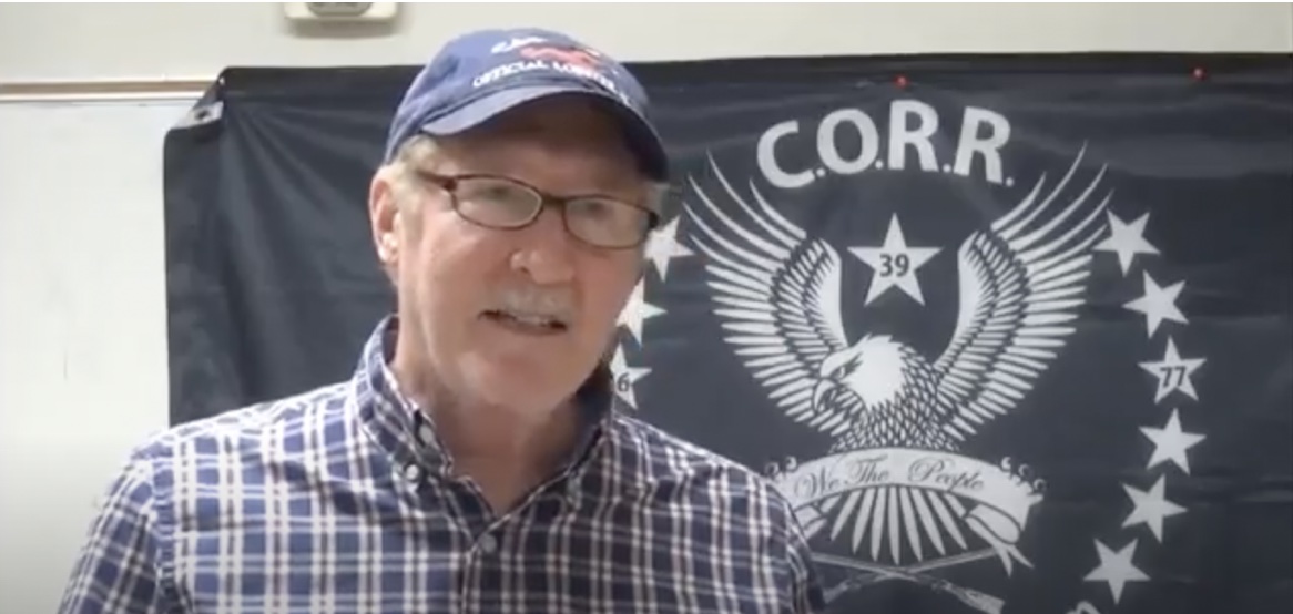 Pastor Mike Johnson, a white-man in his mid-sixties wearing glasses, a checked shirt, and a baseball cap, stands in front of the CORR banner. The CORR banner has an eagle emblem reading "We The People."