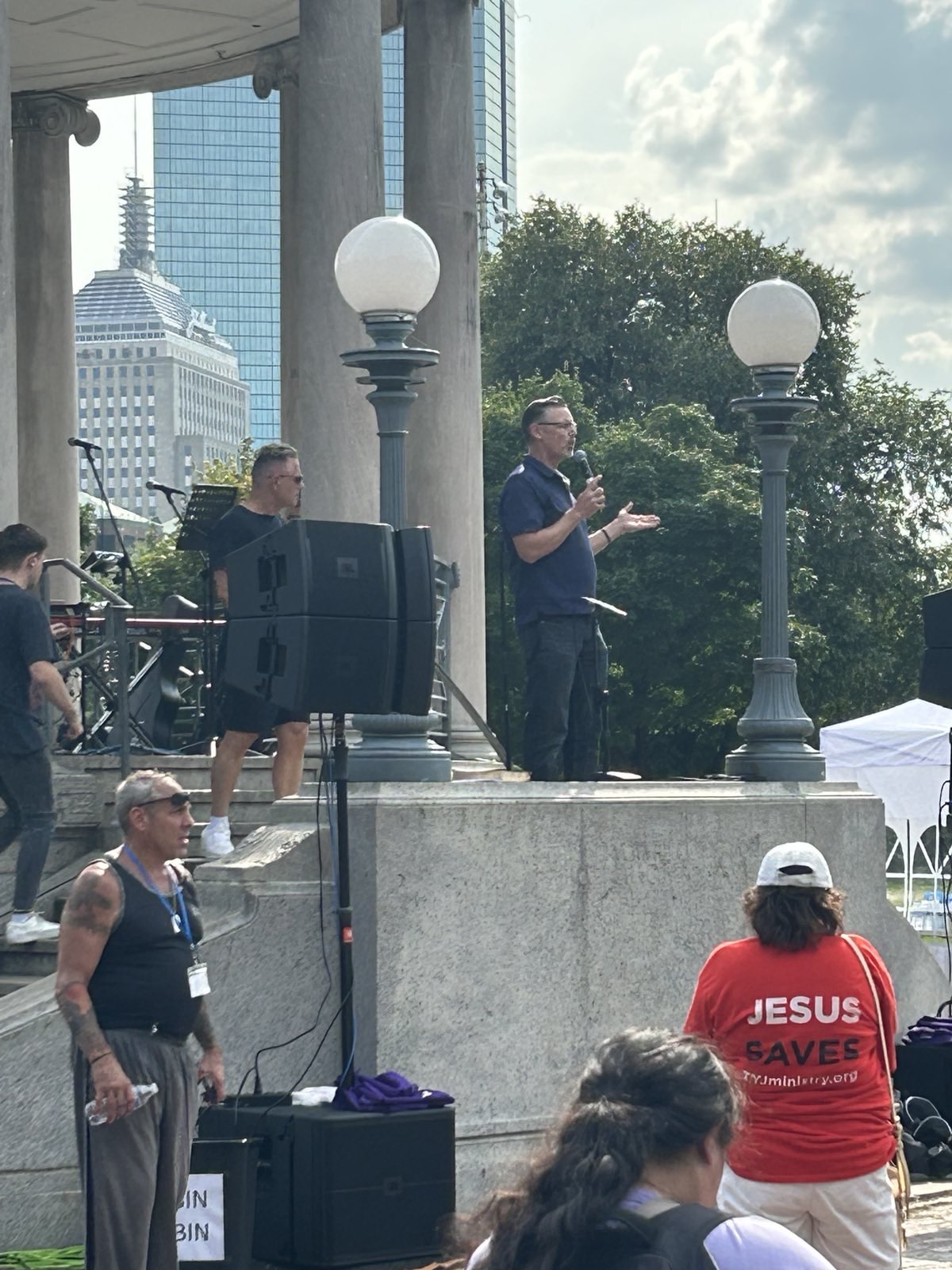 Mike King holds a microphone, addressing the March for Jesus. In the foreground, a listener wears a bright red shirt that reads JESUS SAVES